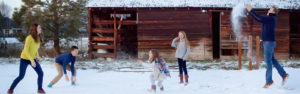 A smiling dad, mom, two young girls and one young boy playing in the snow in front of an old wooden barn.