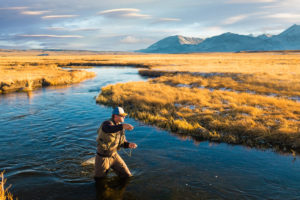 Man standing knee-deep in river with fishing line cast. River is surrounded by flat grass lands with tall snow-capped mountains in the distance.