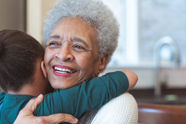 A smiling senior woman with short curly hair giving a big hug to a young child who has his arms wrapped lovingly around her shoulders and neck.