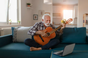 A smiling senior man is sitting cross-legged on a couch in an apartment playing a guitar as he reads music from the screen on a laptop that is sitting on the ottoman that is in front of the couch.