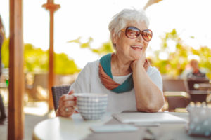 A senior women wearing sun glasses, smiling and holding a large ceramic coffee mug while sitting at a patio table in a wicker chair.