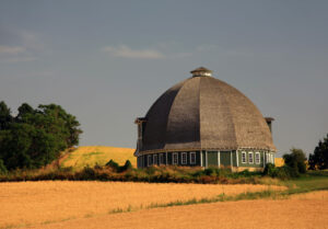 A large dome-shaped barn with windows all around, a cupola and a hay loft. The barn is surrounded by golden wheat fields and grass. Paradise Creek of Olympus Living.